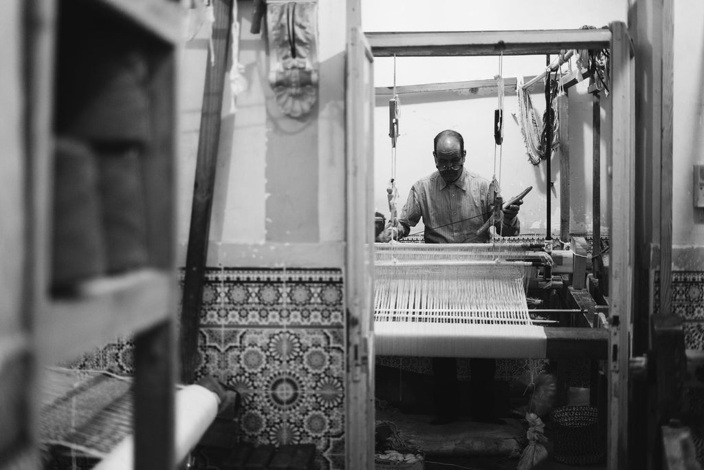 A Moroccan artisan stands at a large loom weaving a blanket in a Moroccan workshop. In the foreground is a wall covered in detailed Moroccan tiles.