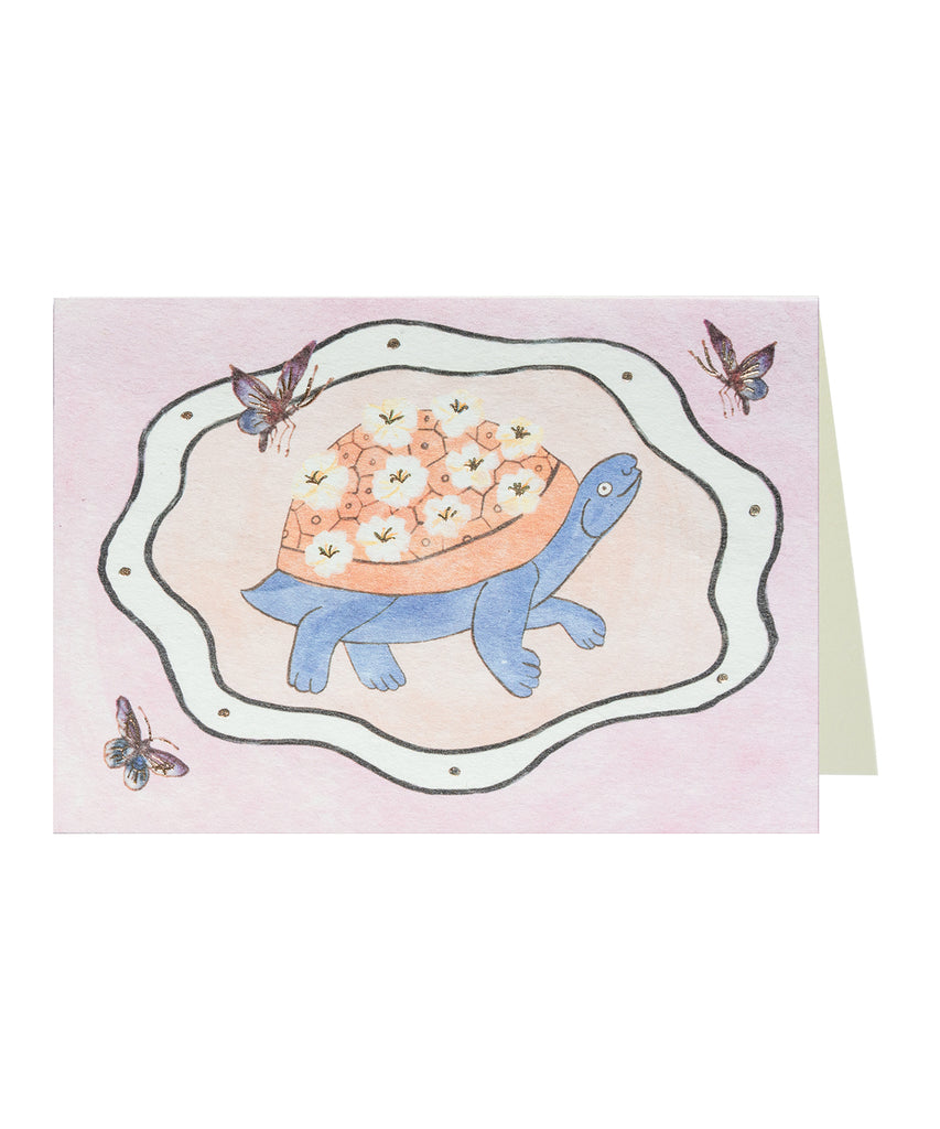 Greeting card and envelope set. Card features a painted illustration of or a blue and orange flowered turtle in the middle of a wavy oval frame with some butterflies flitting around it. Light pink. 
