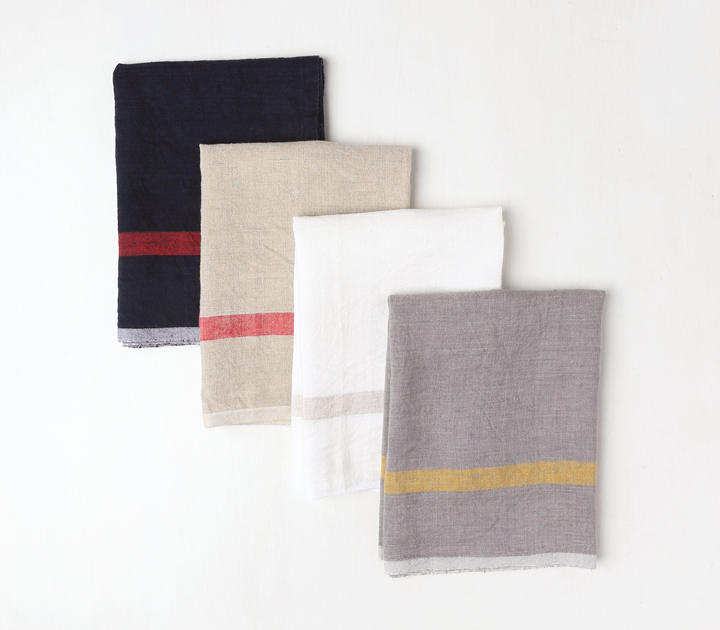 Group of four linen tea towels staggered on top of each other. Black with red stripe in back topped with natural with red stripe topped with white with natural stripe topped with gray with yellow stripe.