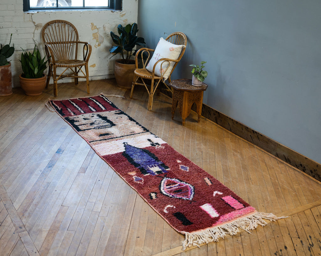 A diagonal view of a vintage Moroccan Boujad runner with a bold modern abstract design in maroon, pink, black, purple, tan, and cream. Various shapes and lines balance each other out in the abstract layout. Rug is surrounded by two rattan chairs, a side table, and potted plants. Wood floors.