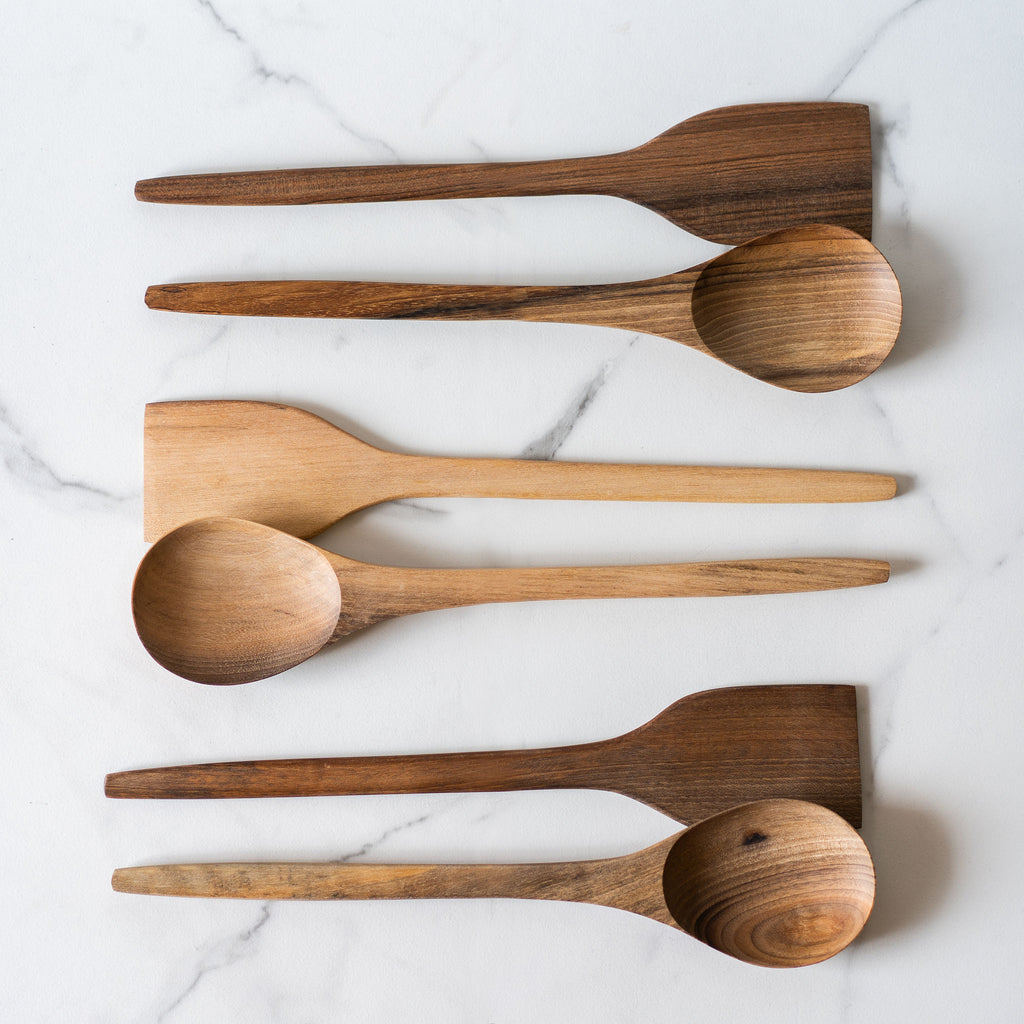 Three spatula and spoon sets in varying tones of hand carved walnut wood lay in a row on a marbled background.