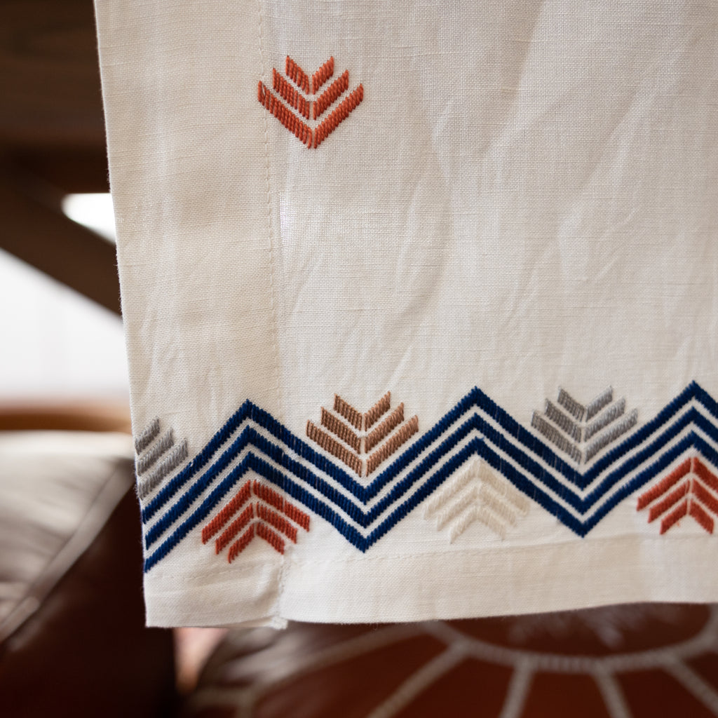 Cream linen table runner with geometric arrow design throughout and zig-zag + arrow combo on edges. Blue, grey, cream, tan, and orange embroidery. Close up of edge embroidery.