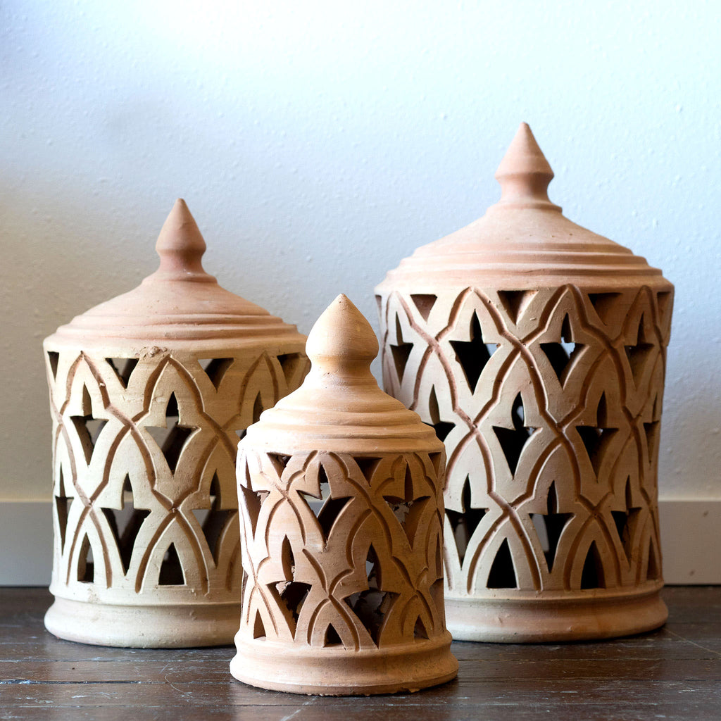 Three sizes of terra cotta lanterns sit on a wood floor in front of a white wall. Traditional punched Moroccan design.