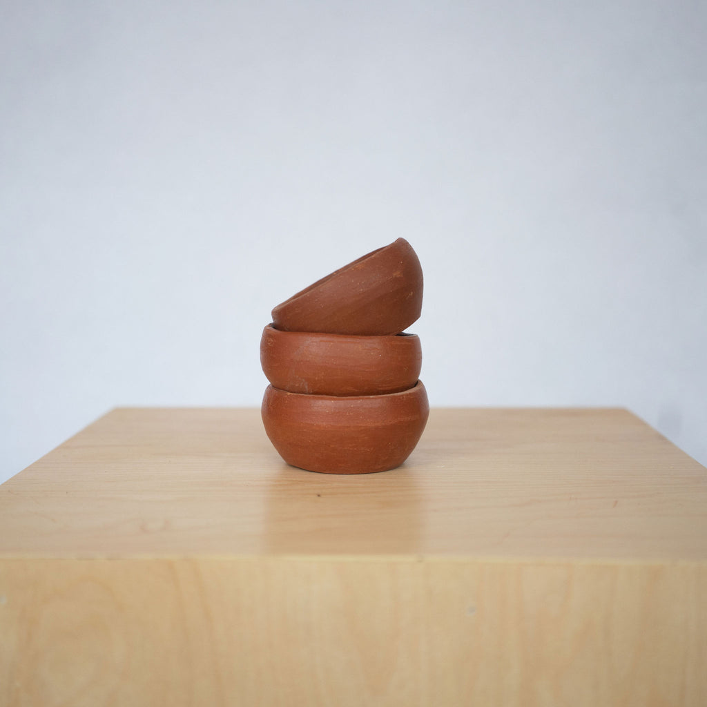 Stack of three small rust toned earthenware bowls sits on a wood platform in front of a white background. 