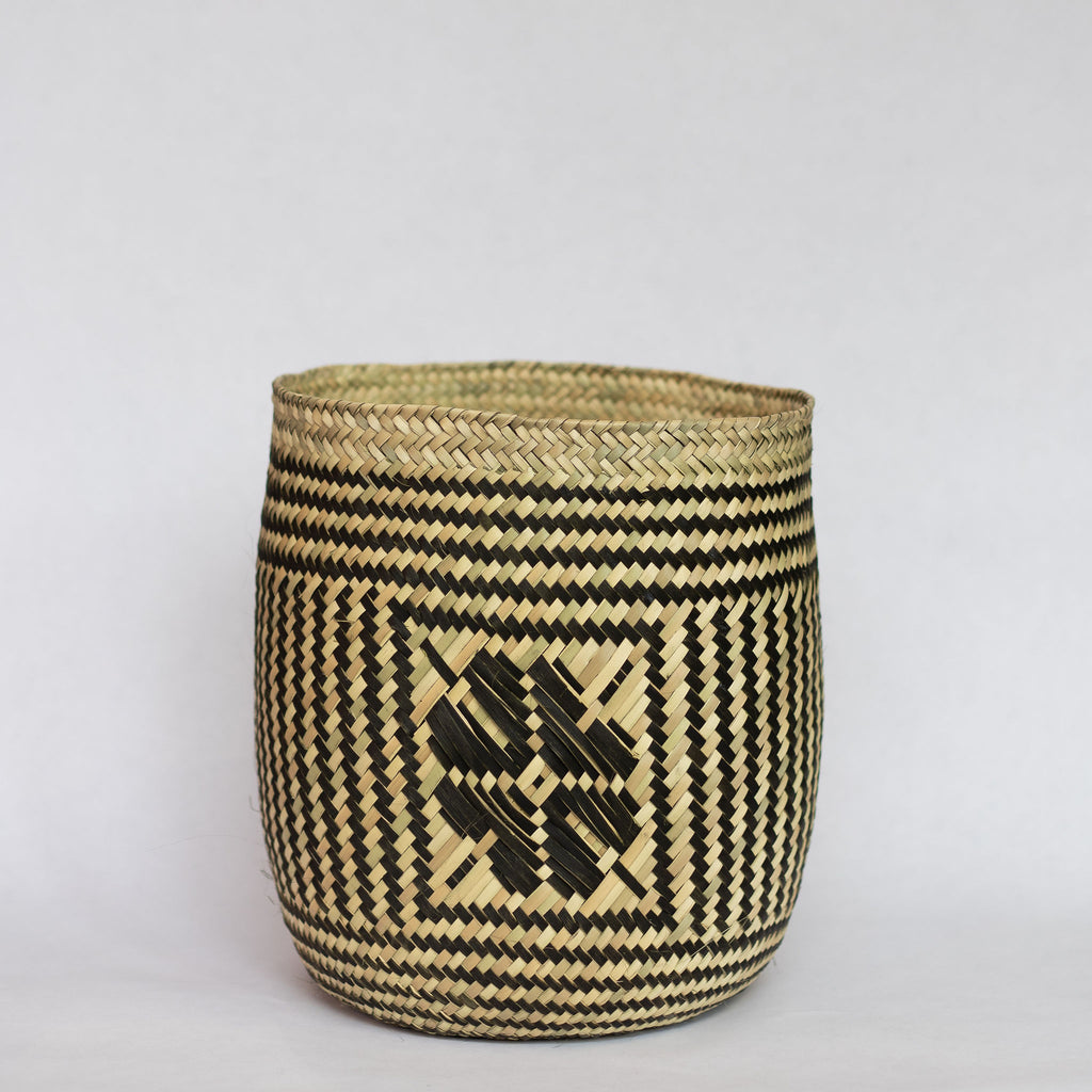 Handwoven palm fiber straight sided basket in traditional Oaxacan designs. Tan and black vertical striped design with flowers on each side. Three black horizontal stripes at top and bottom edge. Gray background.