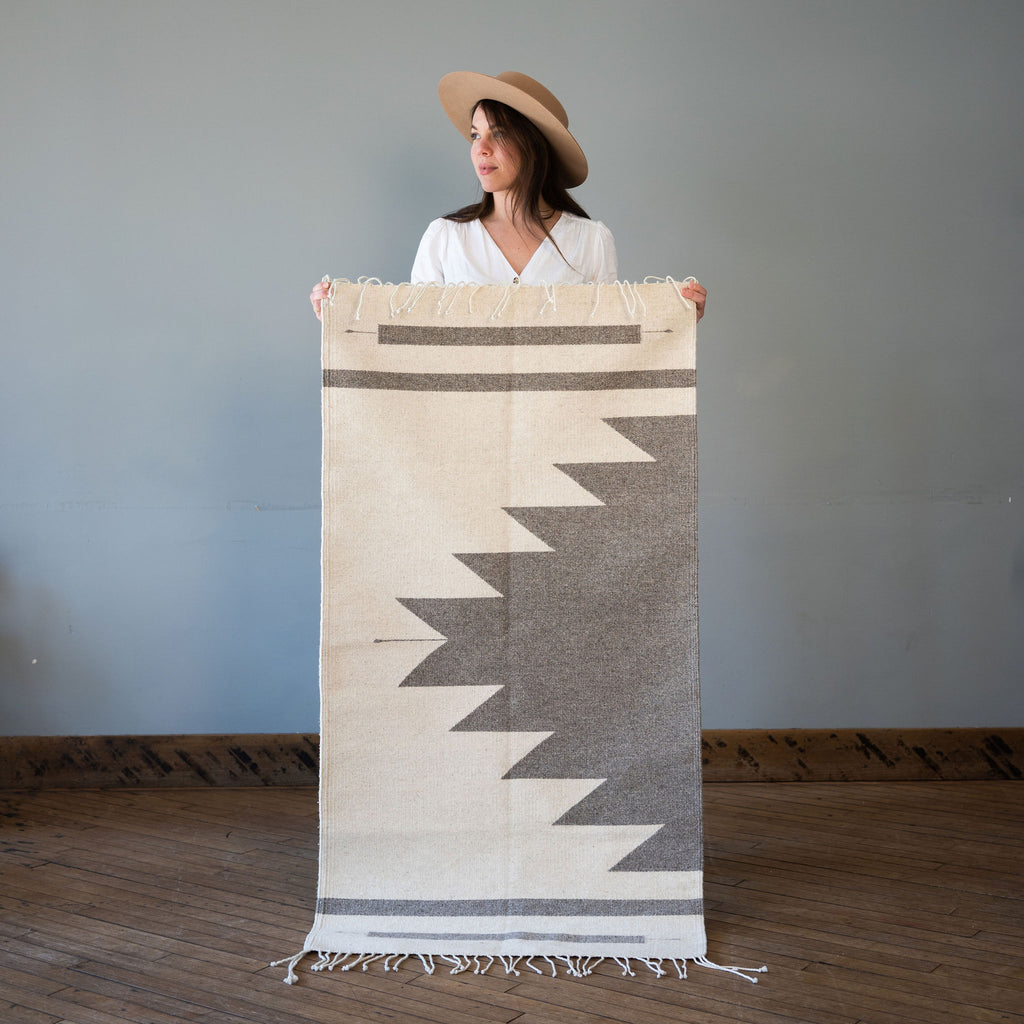 A flat woven wool Oaxacan rug with cream background and gray Aztec-inspired pyramid rising off of one side surrounded by two gray lines on either side. Rug is held up against a grey wall and wood floor by Kelsie in a hat.