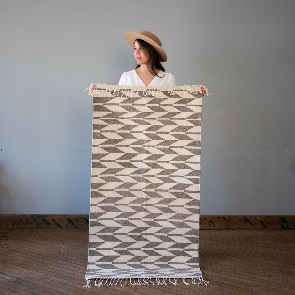 A flat woven cream and light gray Oaxacan rug with a geometric alternating chevron design. The chevron has contrasting lines running though it. Rug is held up against a grey wall and wood floor by Kelsie in a hat.