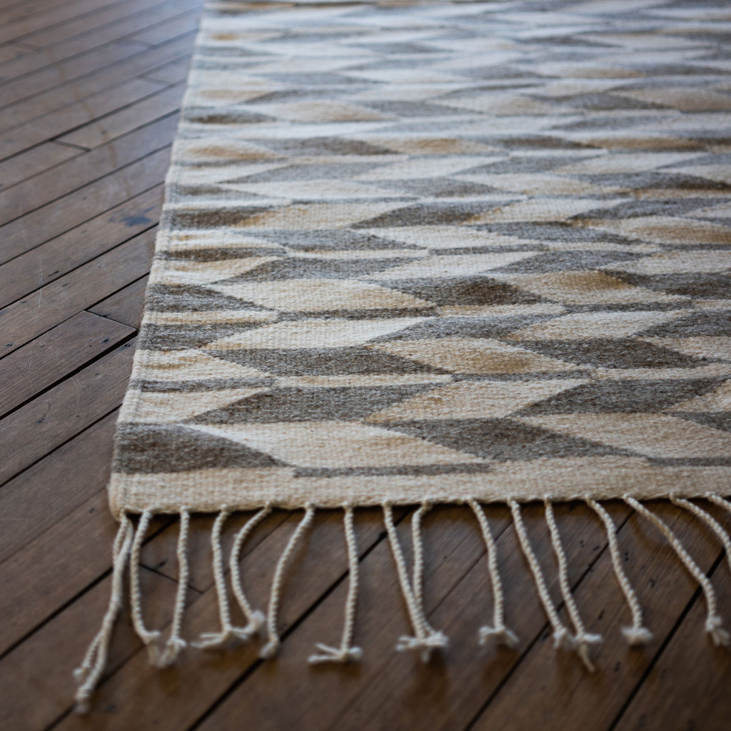 Close up of the corner of a flat woven cream and light gray Oaxacan rug with a geometric alternating chevron design with cream fringe on the edge. Wood floor.