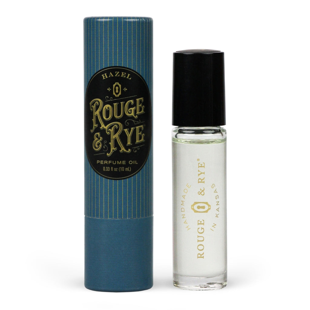 Clear Hazel perfume oil roller in clear glass bottle with black plastic cap next to blue packaging. White background.