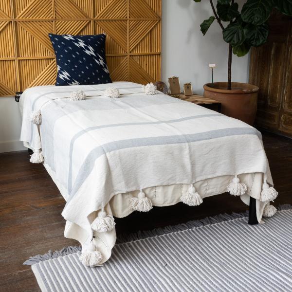 Woven cotton blanket with poms on edge. Natural cream is broken up by varying widths of gray stripes. Blanket is on twin bed in front of a geometric wood headboard.