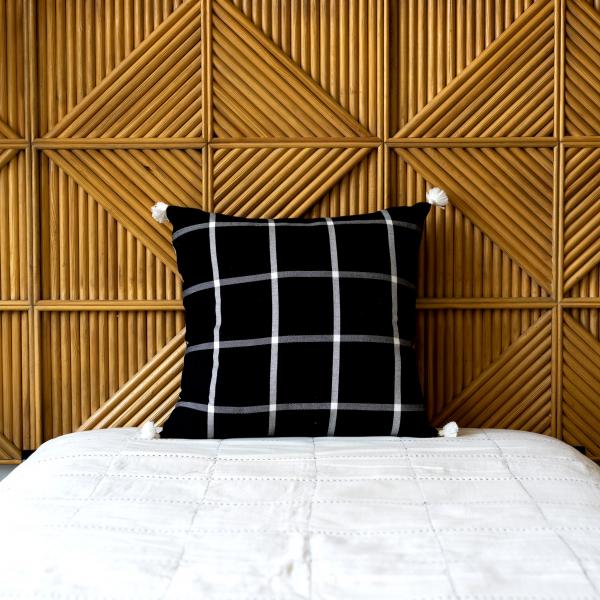 Black woven cotton pillow features a cream grid pattern and cream poms at each corner. Sits on a bed with a white quilt and a wood headboard.