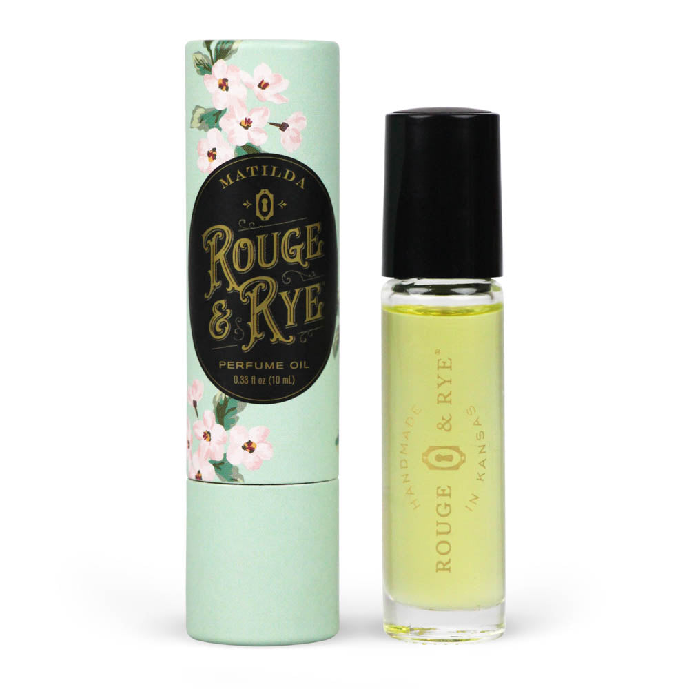 Golden Matilda perfume oil roller in clear glass bottle with black plastic cap next to mint floral packaging. White background.