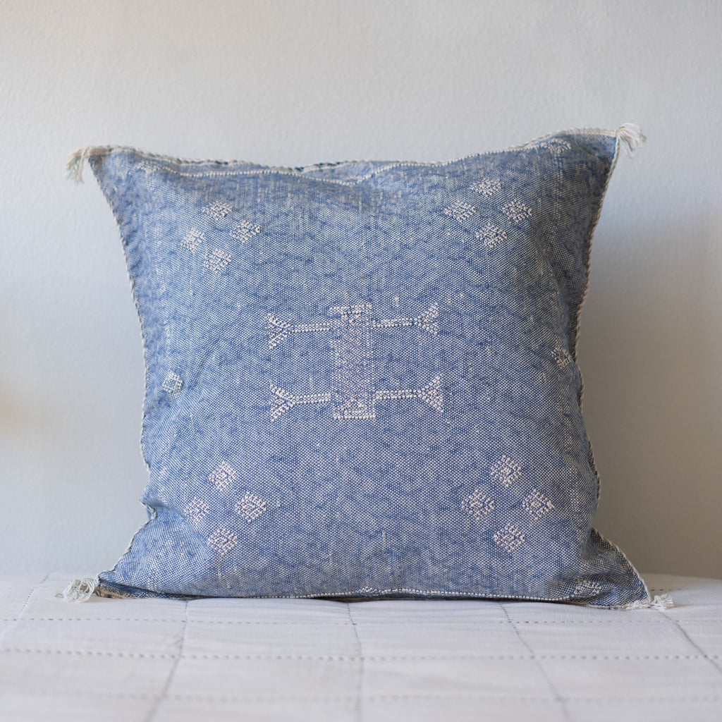 A blue pillow with cream geometric embroidery and small cream tassels on the corners sits on a bed in front of a white wall. 