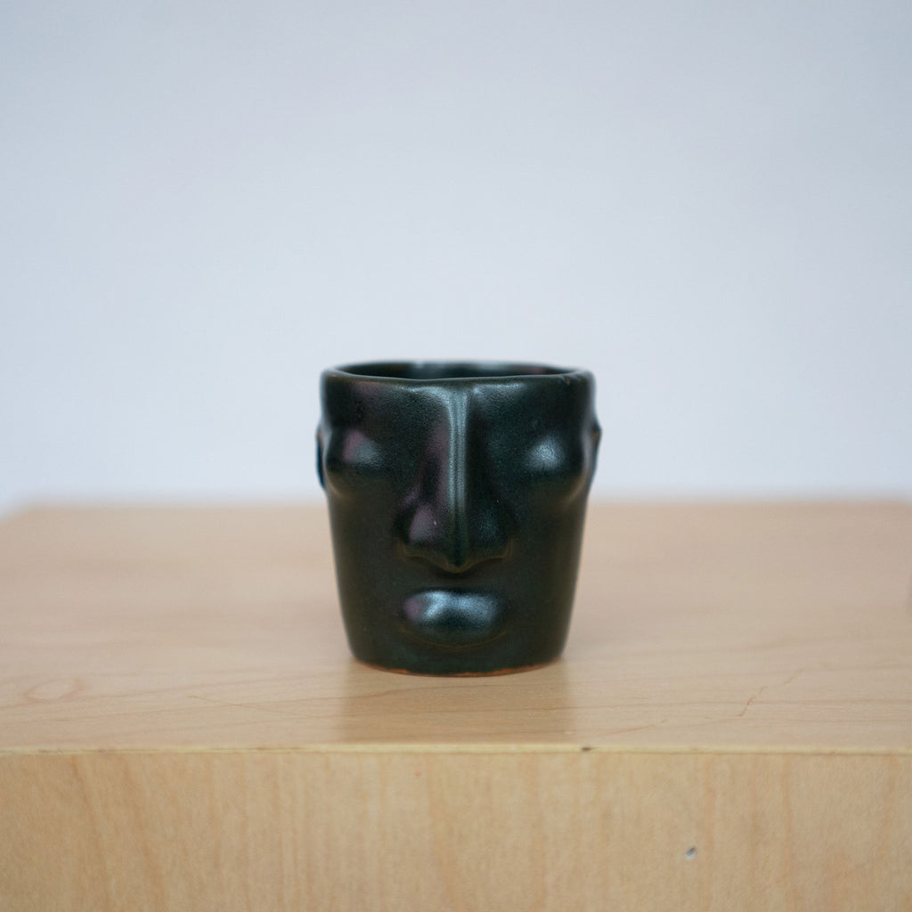 One gloss green ceramic face mezcal cups sit on a wood platform in front of a white background. 