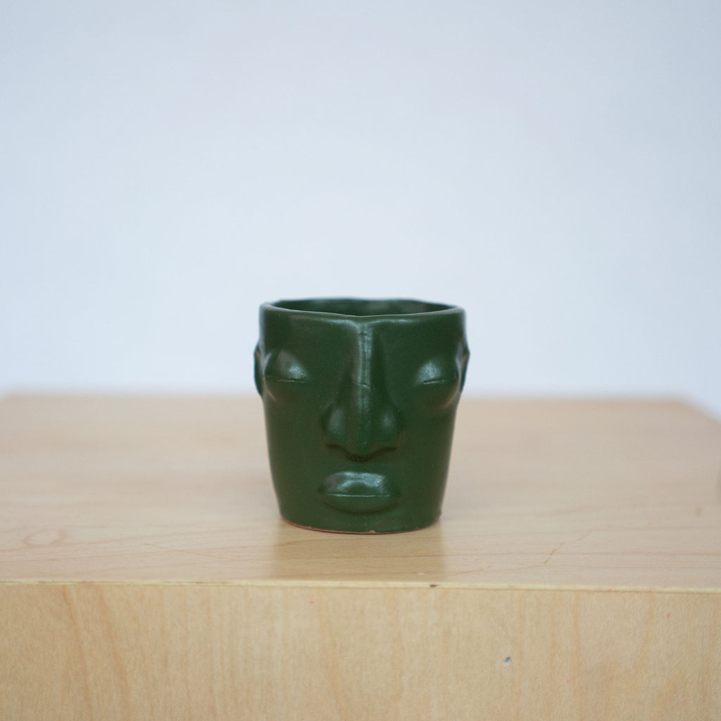 One matte green ceramic face mezcal cups sit on a wood platform in front of a white background. 
