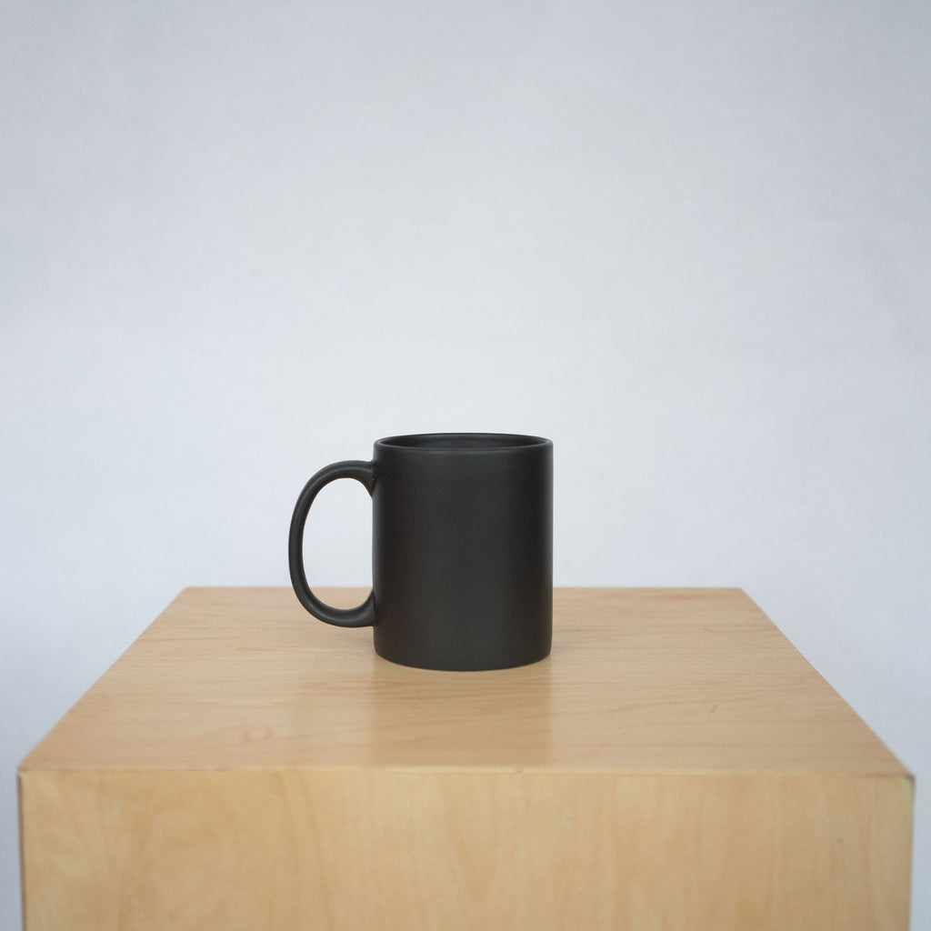 A black ceramic mug with a large handle sits atop a wood pedestal in front of a white background.