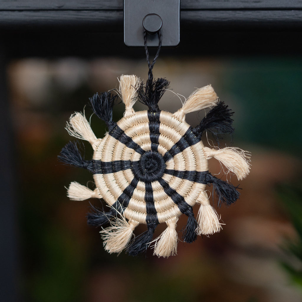Handwoven striped tufted wreath sweetgrass Rwandan tree ornament in tan and black. Blurry shop background.