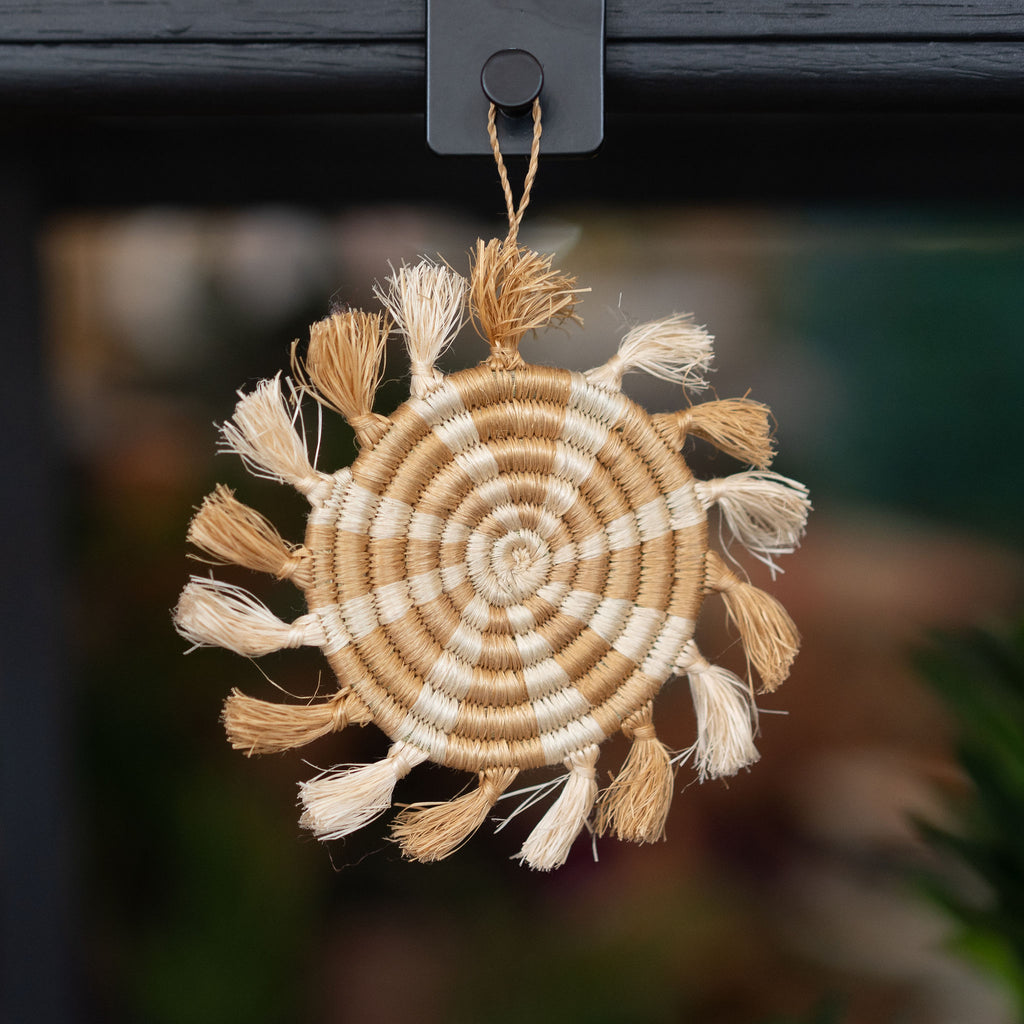 Handwoven striped tufted wreath sweetgrass Rwandan tree ornament in tan and cream. Blurry shop background.