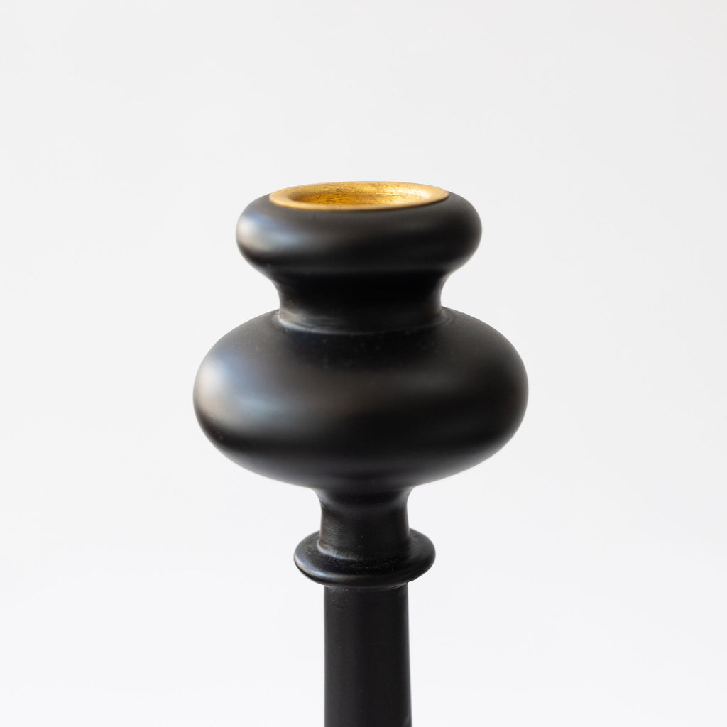 Close up of the gold candle cup at the top of the tall curvy wood taper candlestick with a wide tray at the bottom. Matte black. Sits against a white background.