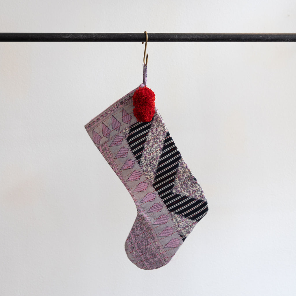 A holiday stocking that is handmade and embroidered with the traditional Kantha stitch. Recycled sari fabric in shades of lavender and black and white stripes join red poms at the hanging loop. Hanging from a brass s-hook from a black bar in front a white background.  