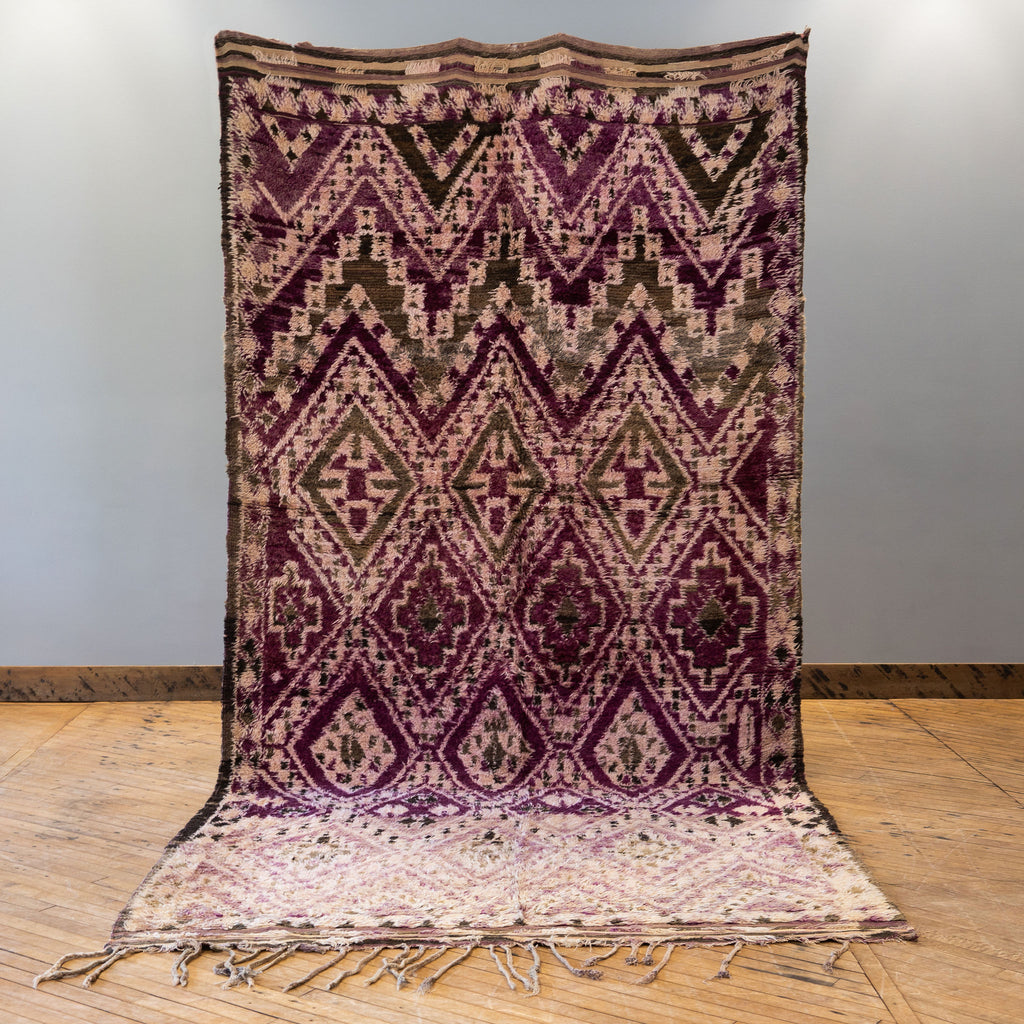 A vintage Moroccan Boujad rug with a bold geometric ombre Berber pattern featuring diamonds and zig zags in shades of purple, olive green, cream, and peach. Rug is held up against a grey wall and wood floor.