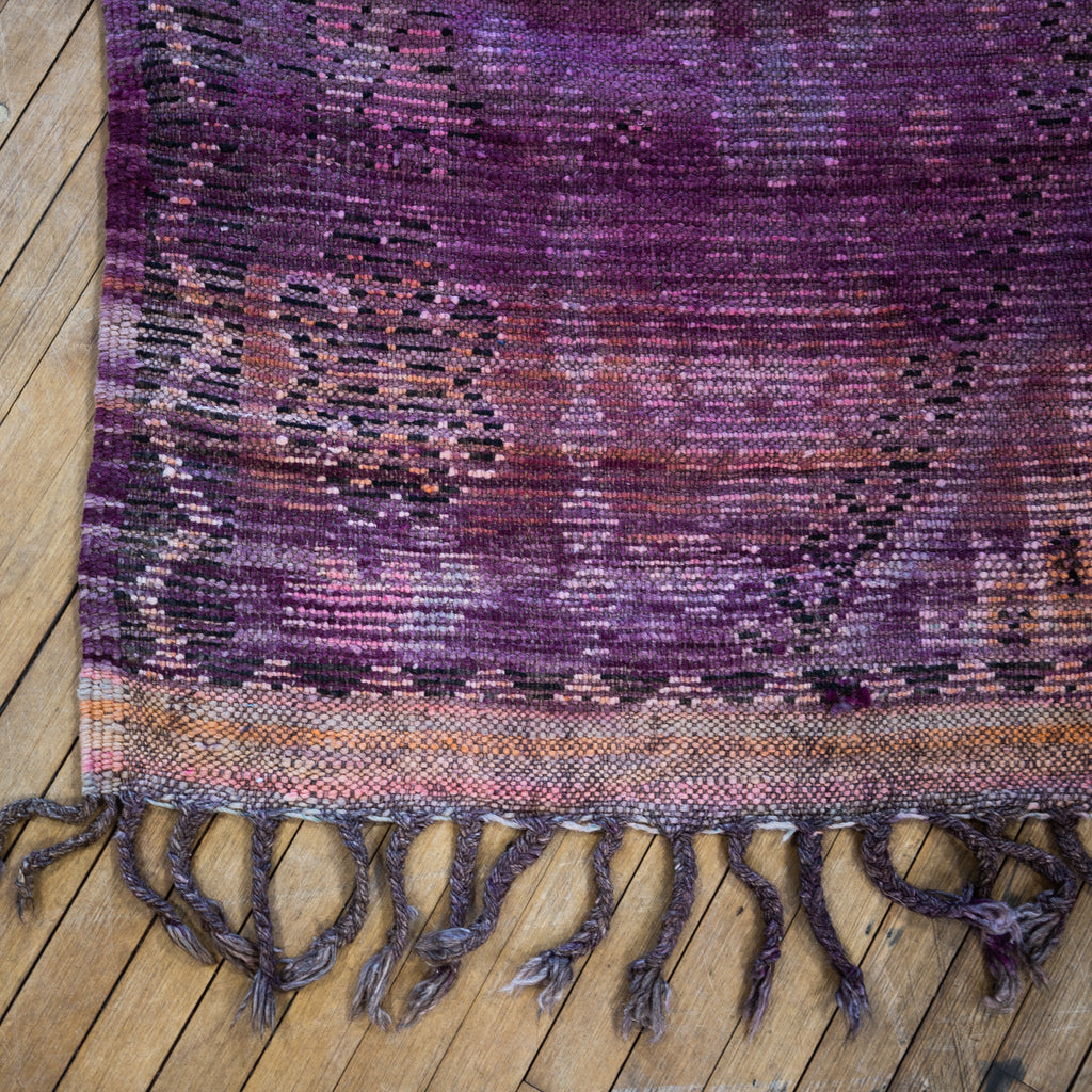Close up of the summer side or back side fringed corner of a vintage Moroccan Boujad rug with a bold diamond design in shades of purple, orange, and pink. Two diamonds are surrounded by various Berber symbols. A zig zag border surrounds an edge section with repeat Berber symbols. Wood floor.