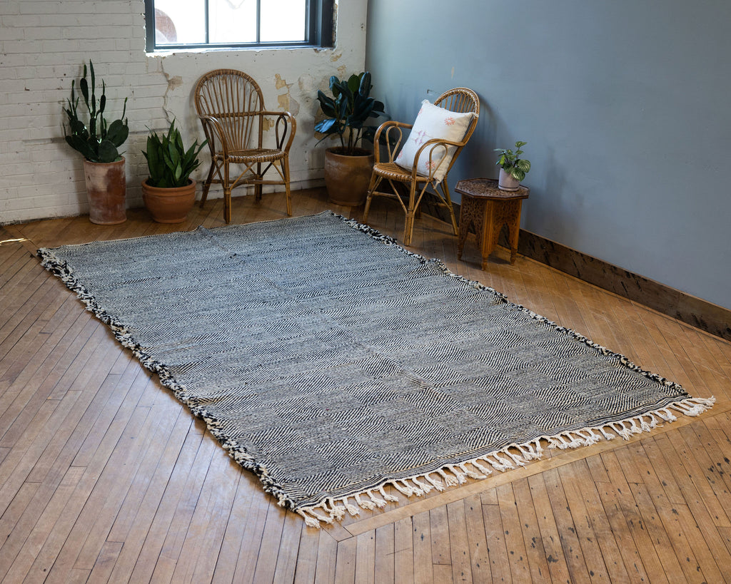 A diagonal view of a flat woven wool Moroccan Zanafi rug with a subtle geometric radiating diamond design in black and white surrounded by two rattan chairs, a side table, and a potted plant. Wood floors.