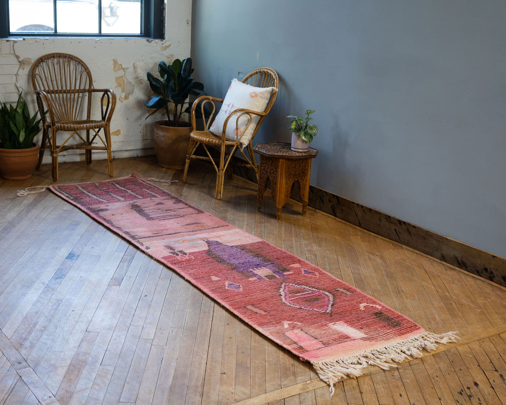 A diagonal view of the back side or summer side of a vintage Moroccan Boujad runner with a bold modern abstract design in maroon, pink, black, purple, tan, and cream. Various shapes and lines balance each other out in the abstract layout. Rug is surrounded by two rattan chairs, a side table, and potted plants. Wood floors.