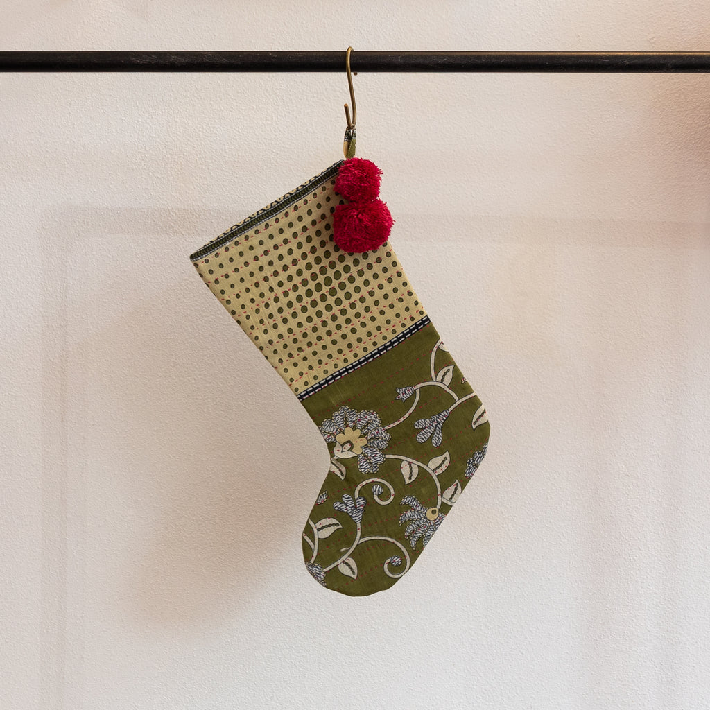 A holiday stocking that is handmade and embroidered with the traditional Kantha stitch. Recycled sari fabric in olive green, pale yellow, blue gray and cream join red poms at the hanging loop. Hanging from a brass s-hook from a black bar in front a white background.
