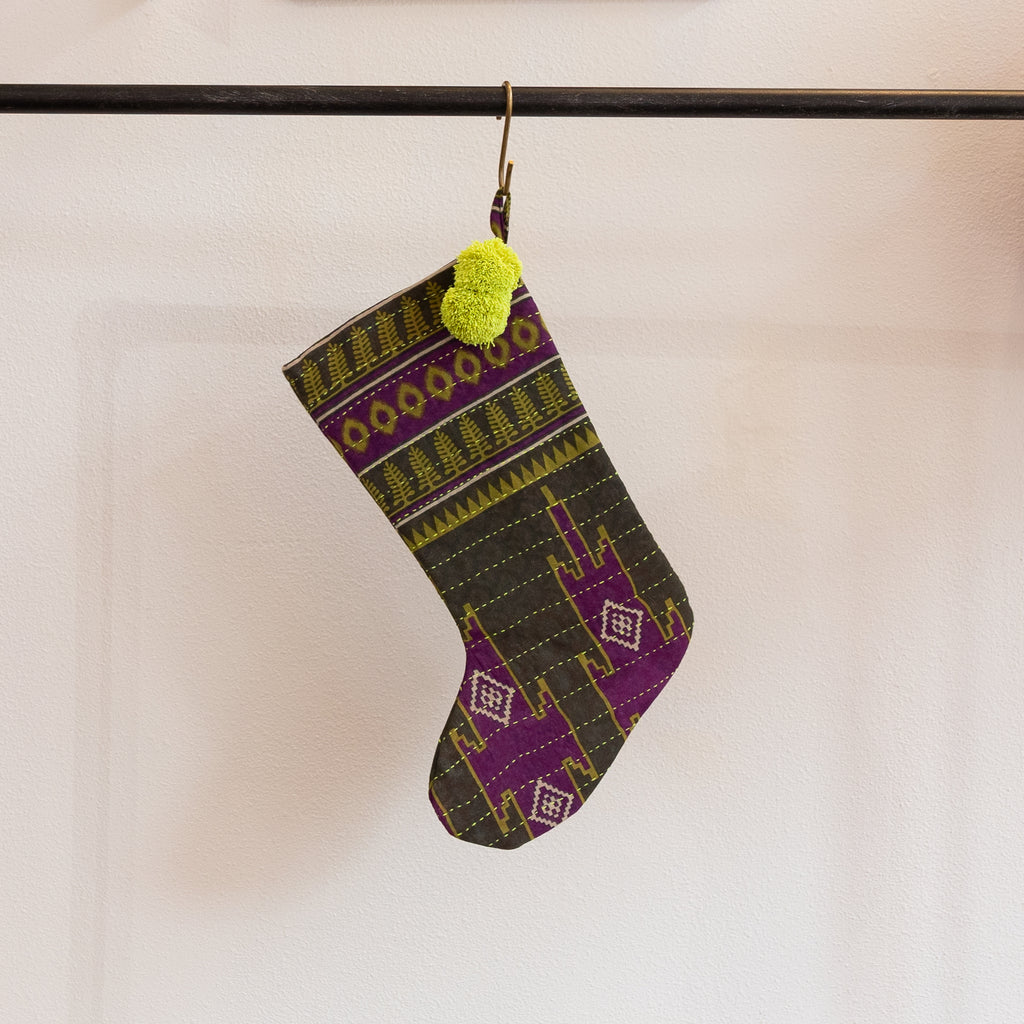 A holiday stocking that is handmade and embroidered with the traditional Kantha stitch. Recycled sari fabric in army green, olive green, orchid purple and cream join bright citrus green poms at the hanging loop. Hanging from a brass s-hook from a black bar in front a white background.