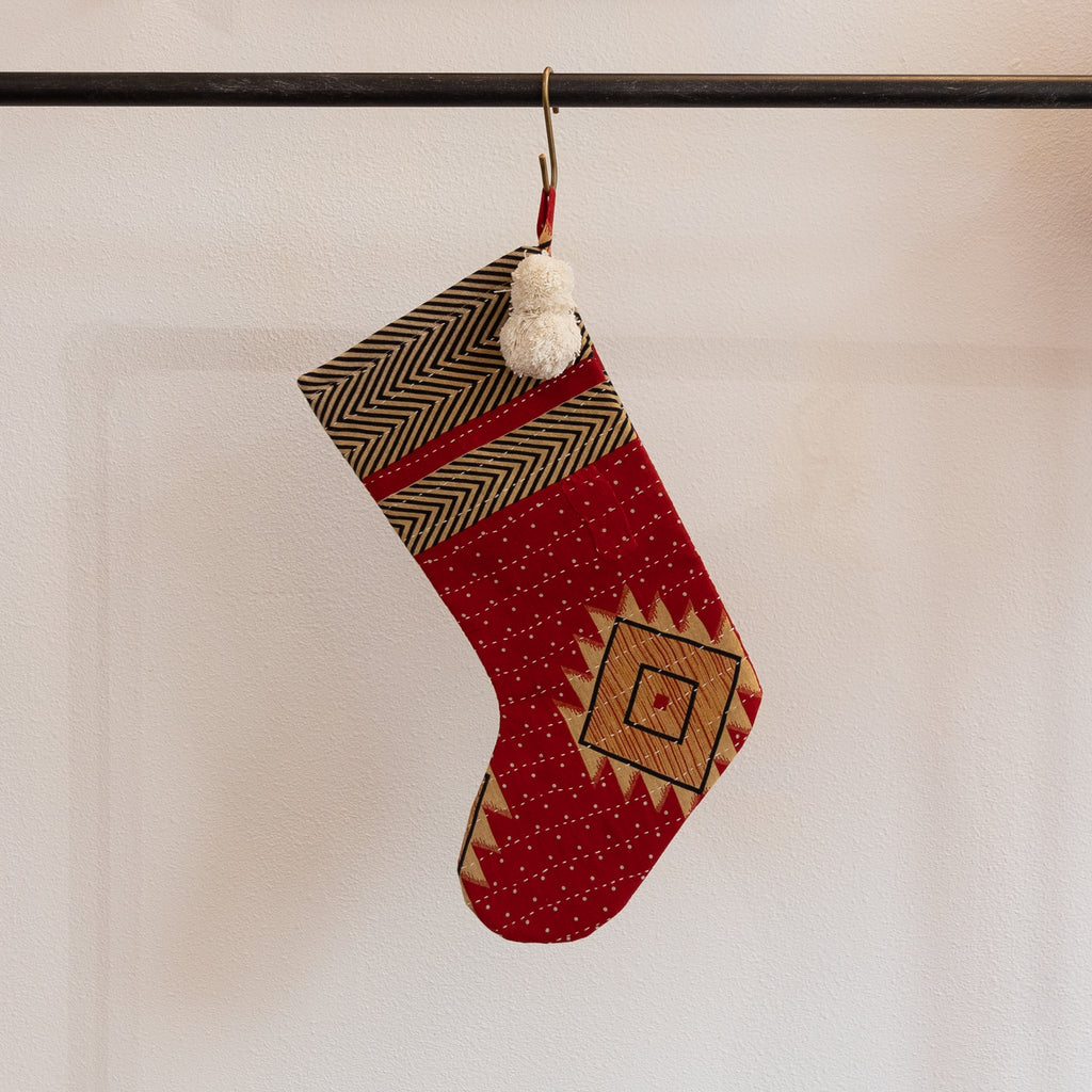 A holiday stocking that is handmade and embroidered with the traditional Kantha stitch. Recycled sari fabric in red, black, pale yellow and cream join cream poms at the hanging loop. Hanging from a brass s-hook from a black bar in front a white background.