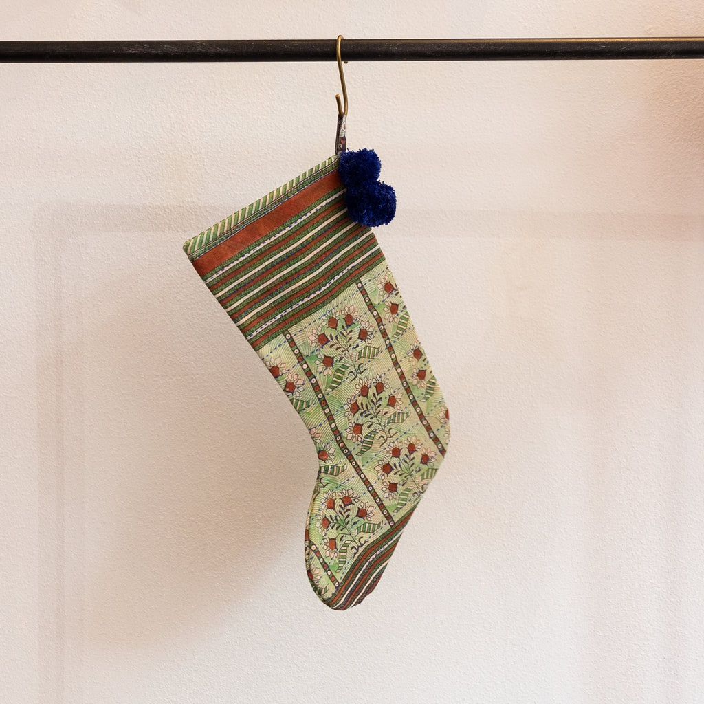 A holiday stocking that is handmade and embroidered with the traditional Kantha stitch. Recycled sari fabric in rust red, shades of green, and cream join navy blue poms at the hanging loop. Hanging from a brass s-hook from a black bar in front a white background.