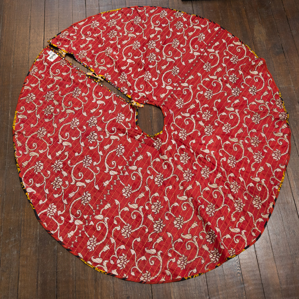 A tree skirt that is handmade and embroidered with the traditional Kantha stitch over recycled sari fabric in primarily cream and red with yellow accents. Reversible with one side featuring a floral pattern and the other side a zig zag pattern. 