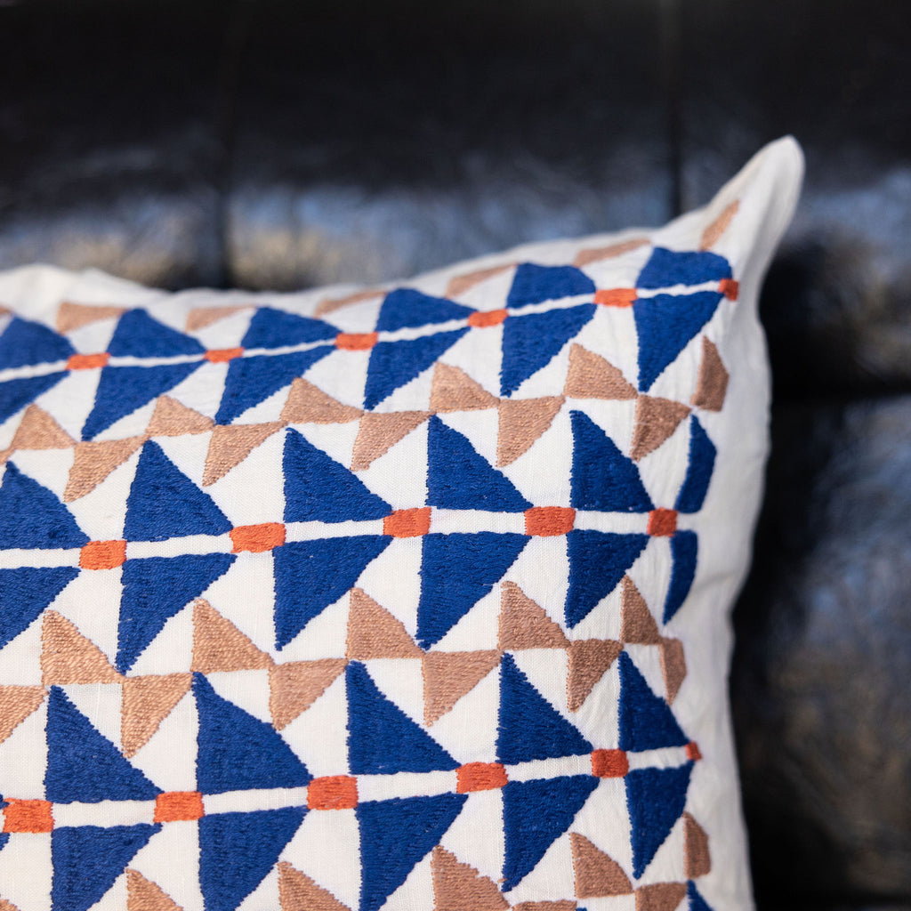 Square pillow featuring Puktadozi style embroidery with blue + tan triangles and orange dashes. Close up of design.