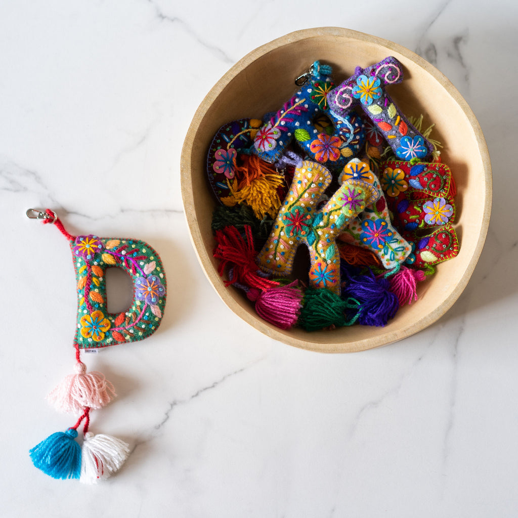 A wood bowl of various wool letter keychains embroidered in a bright Peruvian floral style. Each letter has a pom hanging at the bottom and a clip at the top. A 'D' keychain sits next to the bowl on a marble background.