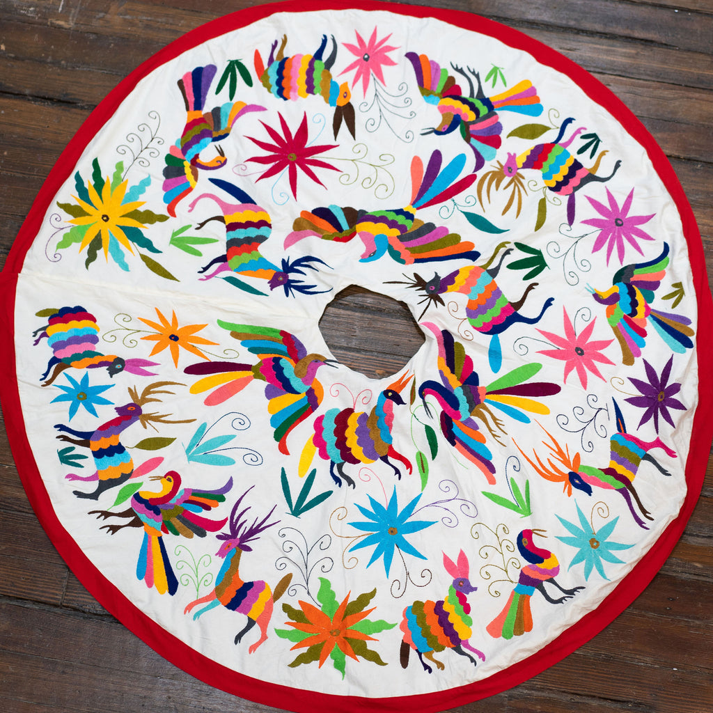 Christmas tree skirt in traditional Otomi embroidered style with bright multi-colored flora and fauna on a cream background with red border. Spread out on a wood floor