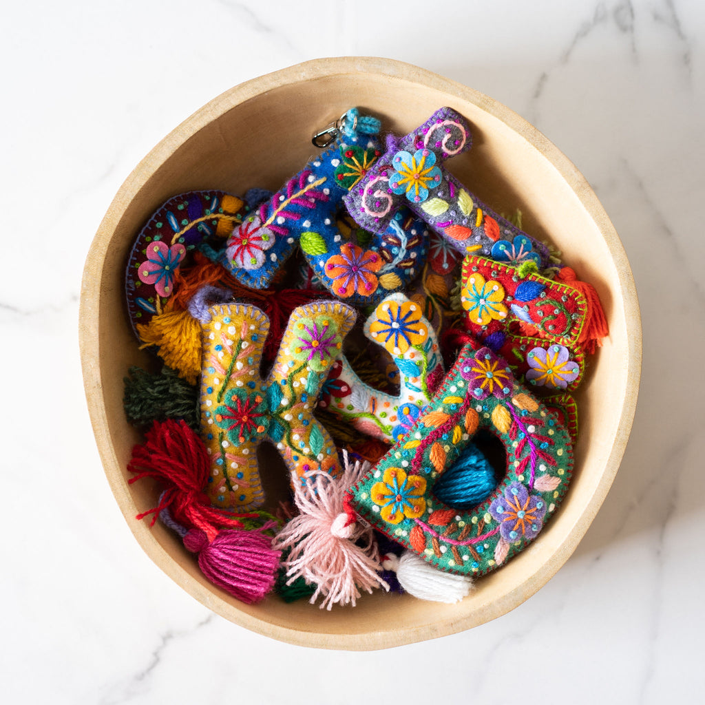 A wood bowl of various wool letter keychains embroidered in a bright Peruvian floral style. Each letter has a pom hanging at the bottom and a clip at the top.