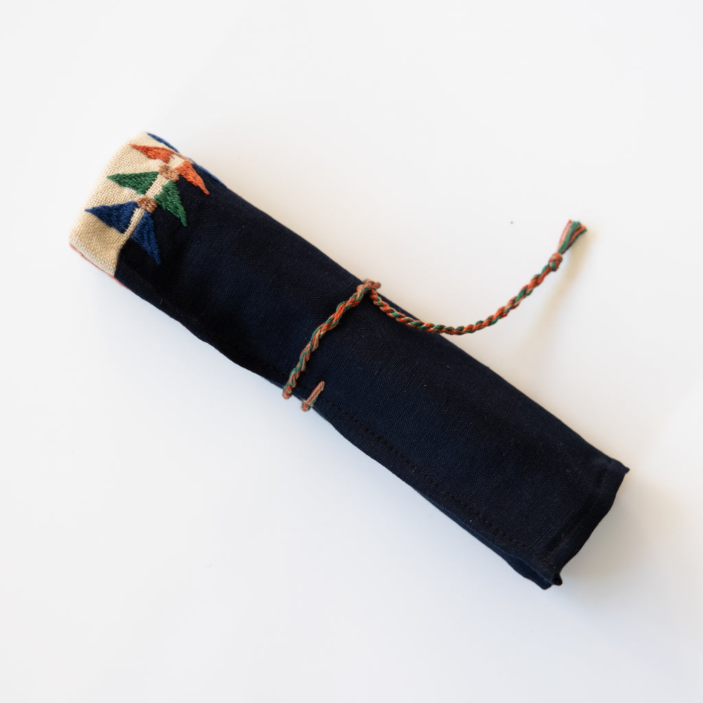 Embroidered roll up pencil case. Black and tan with blue,  orange, green, tan embroidery. Shown rolled up and tied. Triangle embroidery runs along top edge.