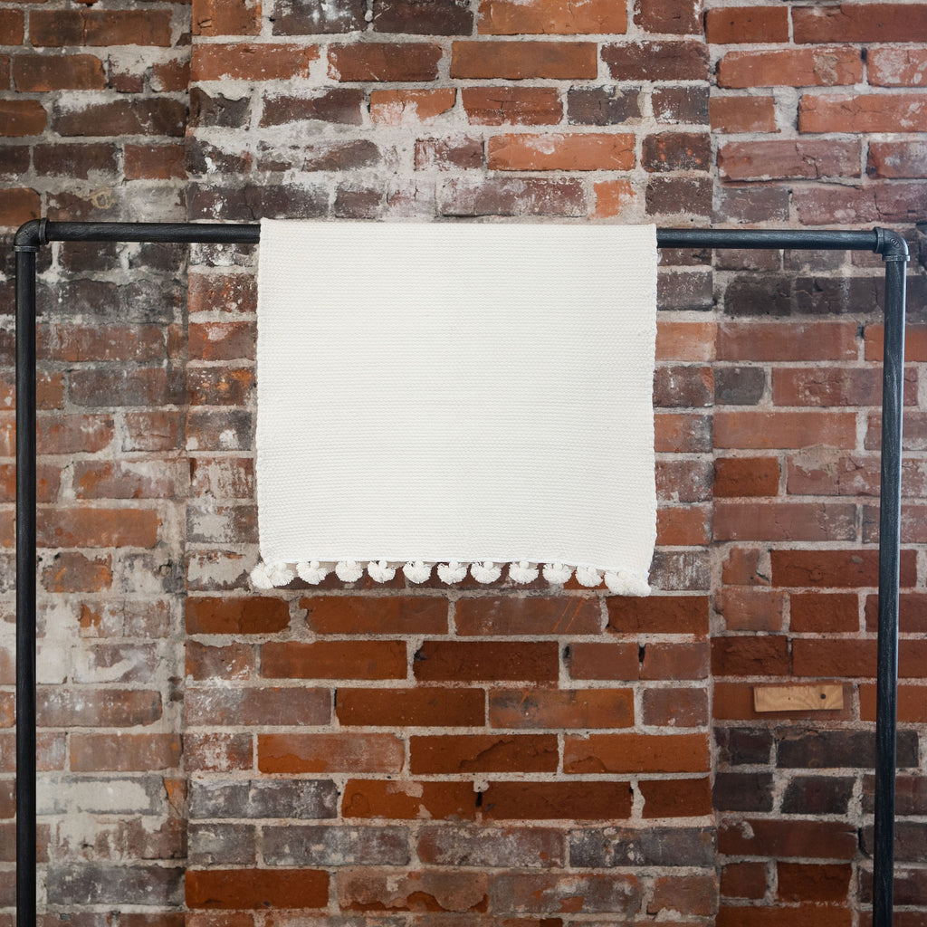 An organic cotton white woven bath mat with tasseled edges hangs over a black rod in front of a brick wall. 