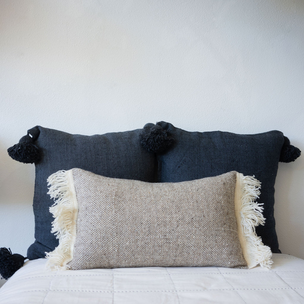 Tan and cream wool tween lumbar pillow in front of two navy cotton pillows with poms on the corners.