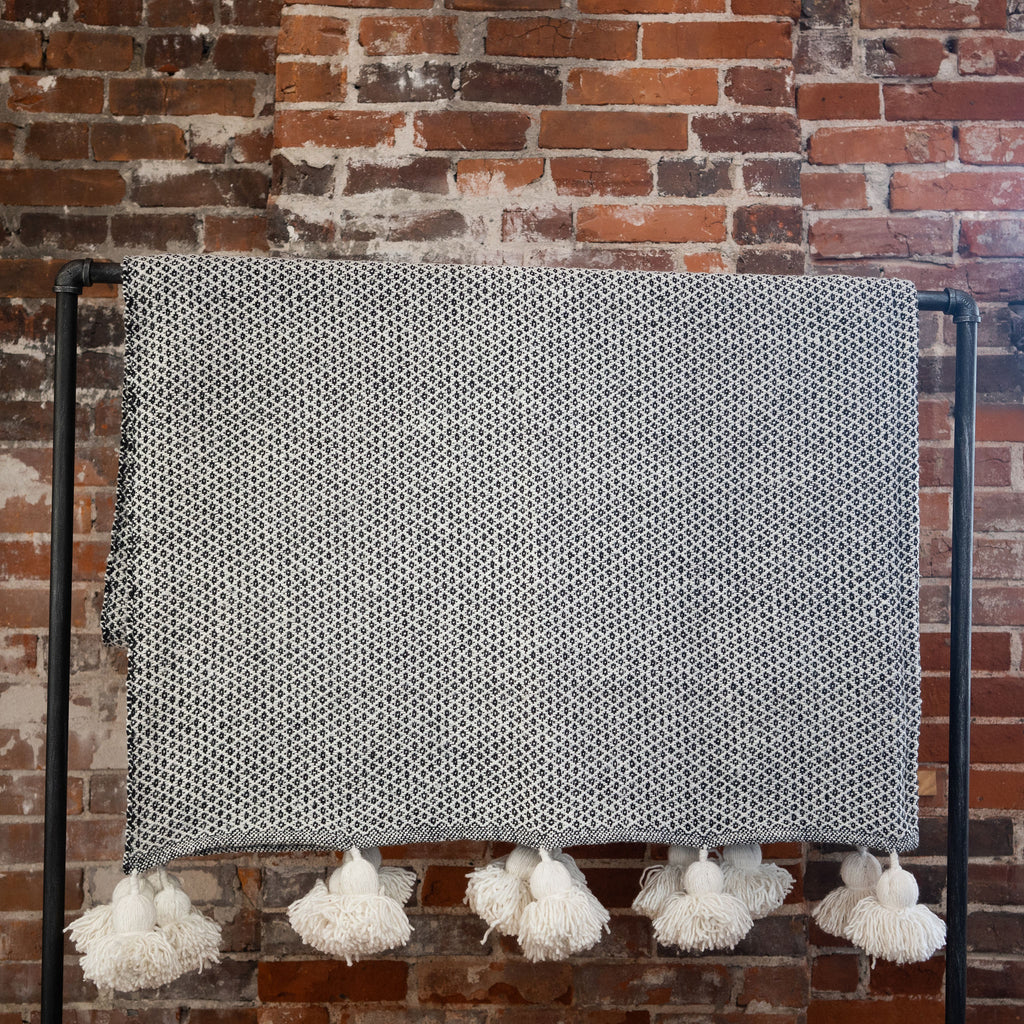 Large cotton blanket woven in a black and white small diamond pattern with big white pom poms along the edge.