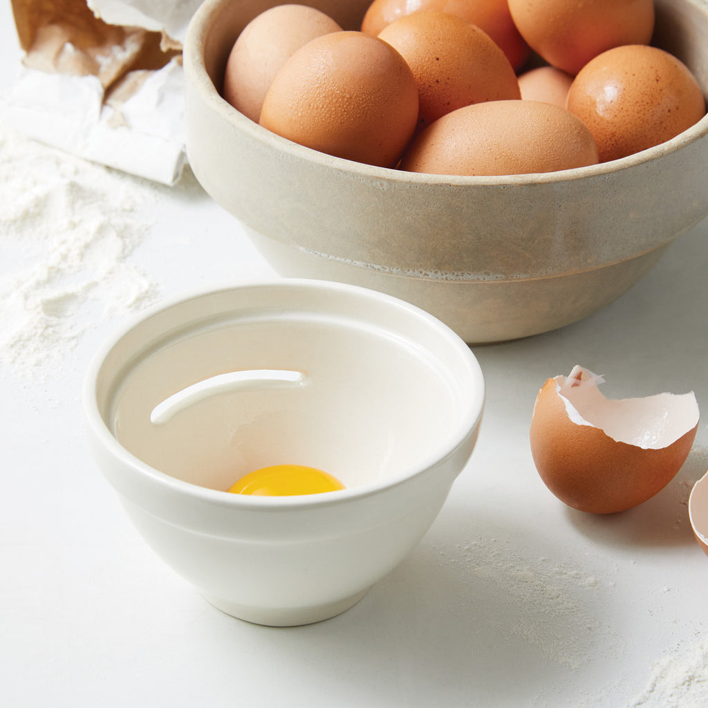 Cream colored ceramic egg separator with an egg inside next to a broken egg shell and bowl of eggs.
