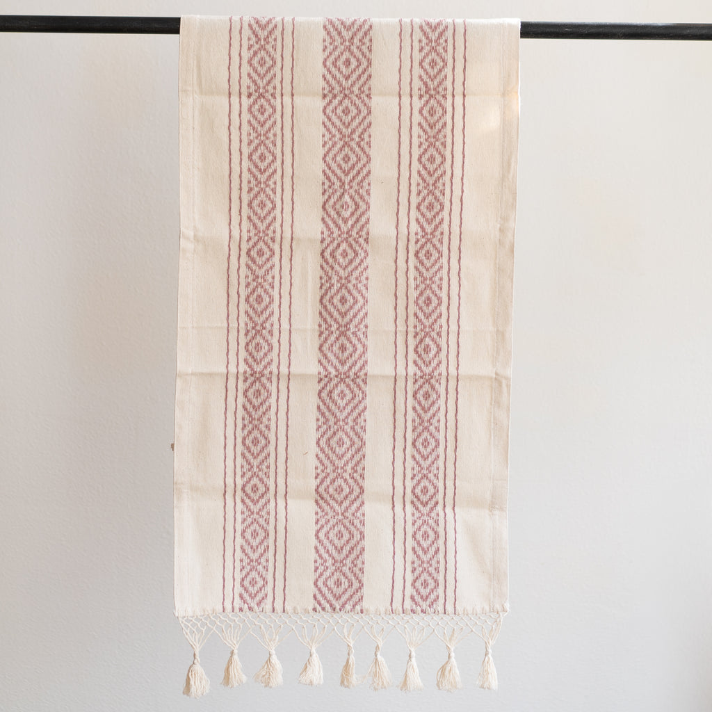 Handwoven cotton table runner in traditional Oaxacan design. Rose colored radiating diamonds on three stripes running the long way on cream background. Macrame ending in cream colored knots and tassels on the edge.