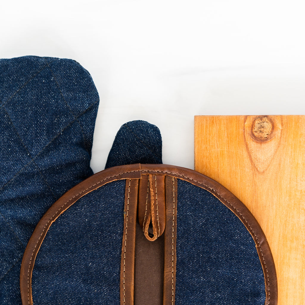 Close up of a leather + denim pot holder + oven mitt combo laying near a cutting board.