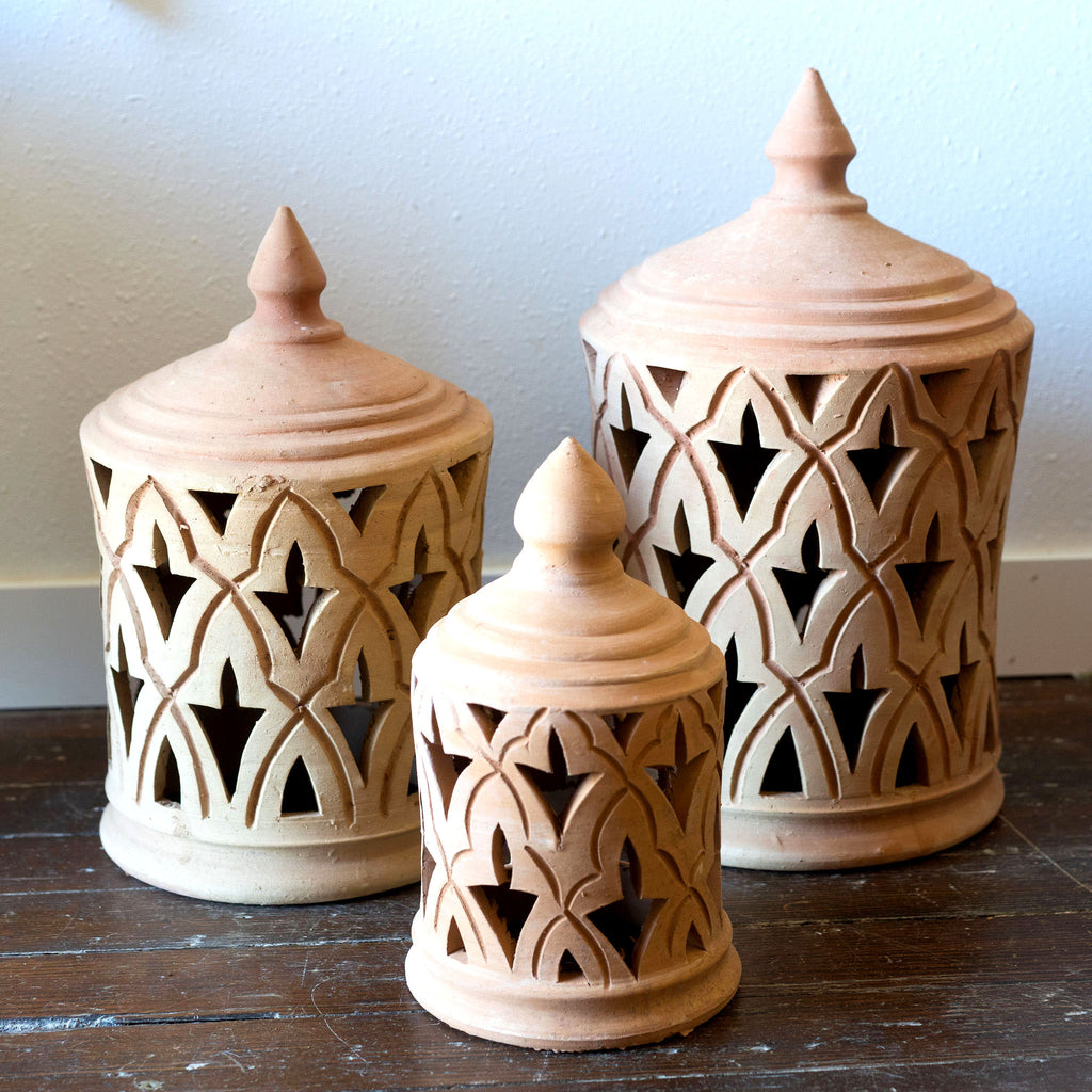 Three sizes of terra cotta lanterns sit on a wood floor in front of a white wall. Traditional punched Moroccan design.