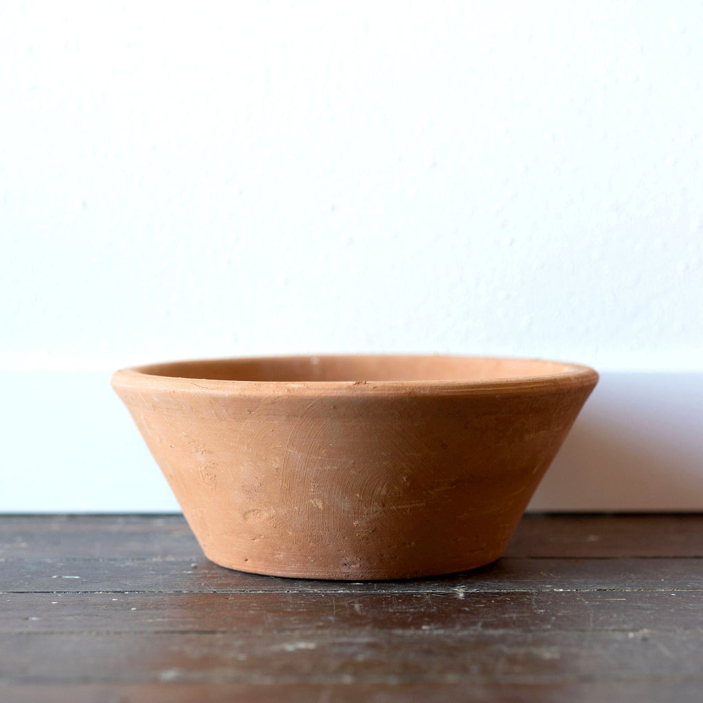 A low round terra cotta pot on wood floor in front of white wall.