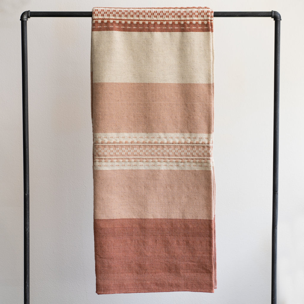 Naturally dyed wool blanket in tones of terra cotta and cream with varying widths of color blocking, stripes, and traditional Oaxacan weaving techniques. Hangs over black bar in front of white background.