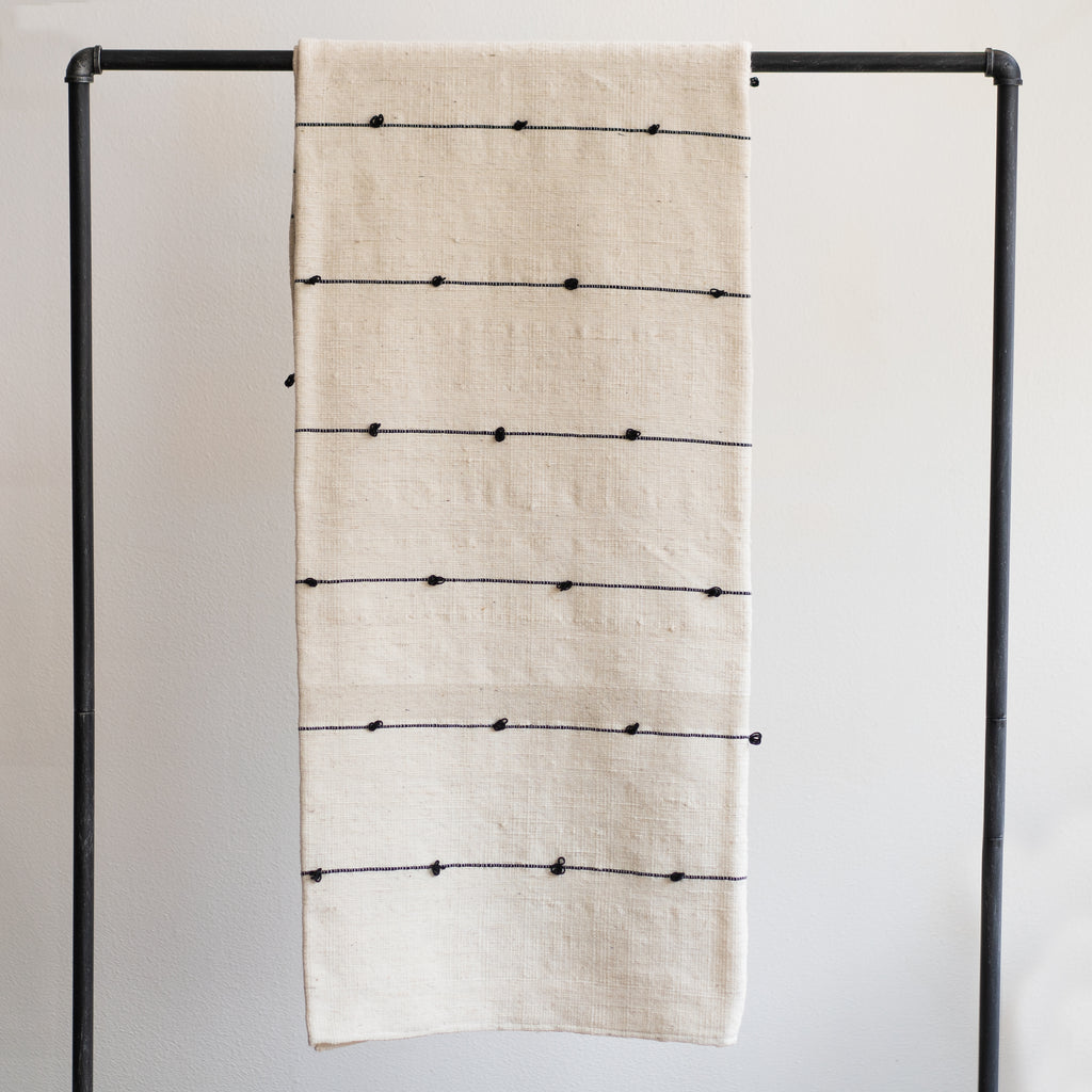Naturally dyed wool blanket with black tufted lines on cream background. Hangs over black rod in front of white background. 