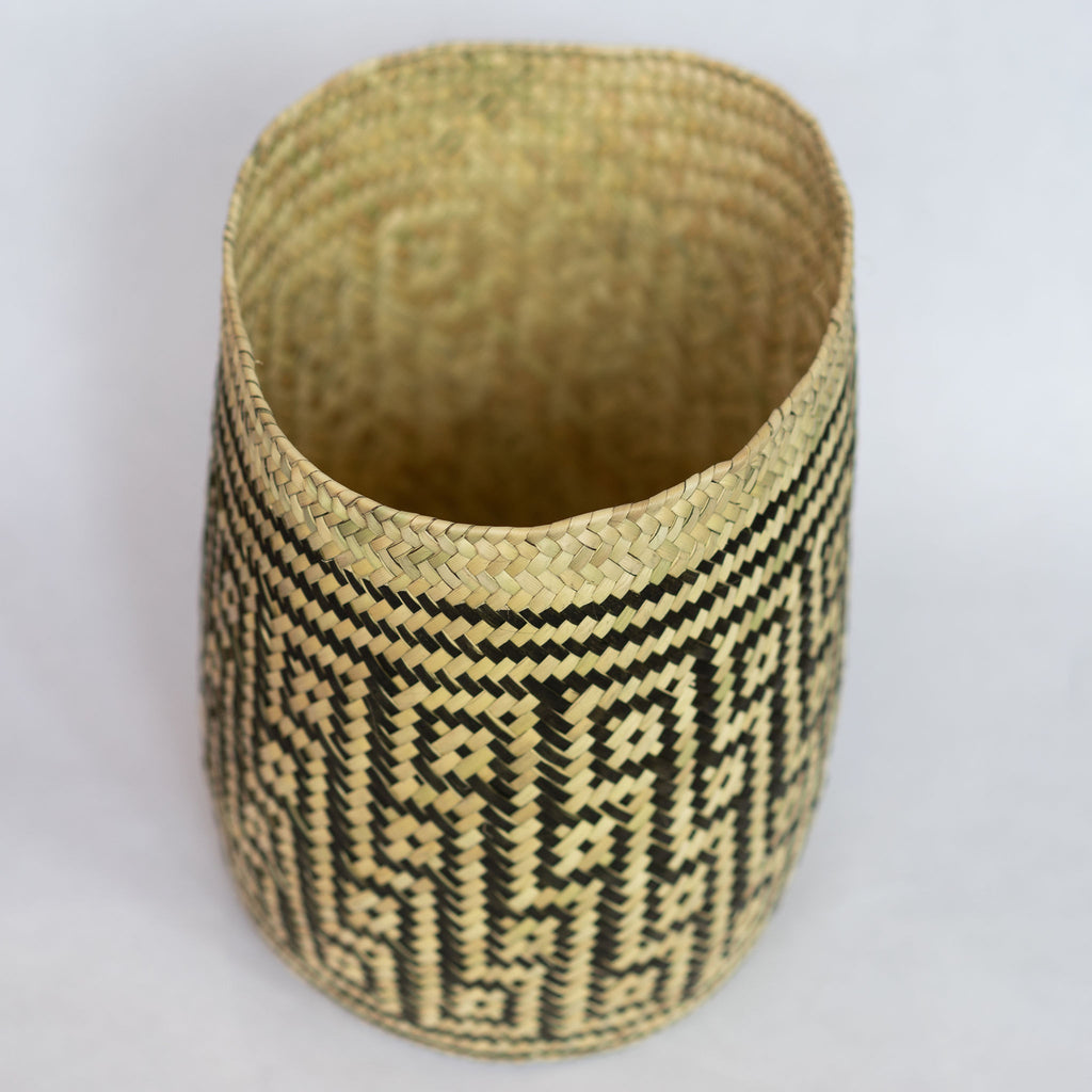 Handwoven palm fiber straight sided basket in traditional Oaxacan designs. Tan and black maze design with squares in diagonal rows with contrasting dots inside each square. Three black stripes at top and bottom edge. Inside is natural tan. Gray background.