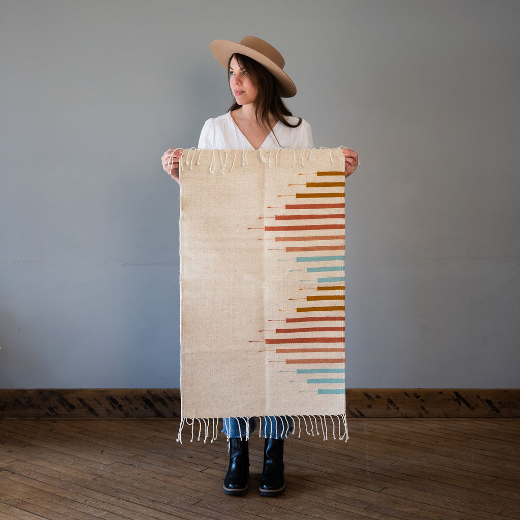 A flat woven cream Oaxacan rug with a geometric triangle made out of lines in Light Blue, Blush, Terracotta, Ochre. Rug is held up against a grey wall and wood floor by Kelsie in a hat.
